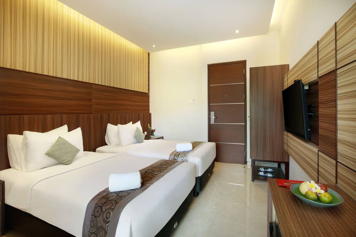 A hotel room with two beds, a TV, a small table with fruit, and a wooden panel wall.
