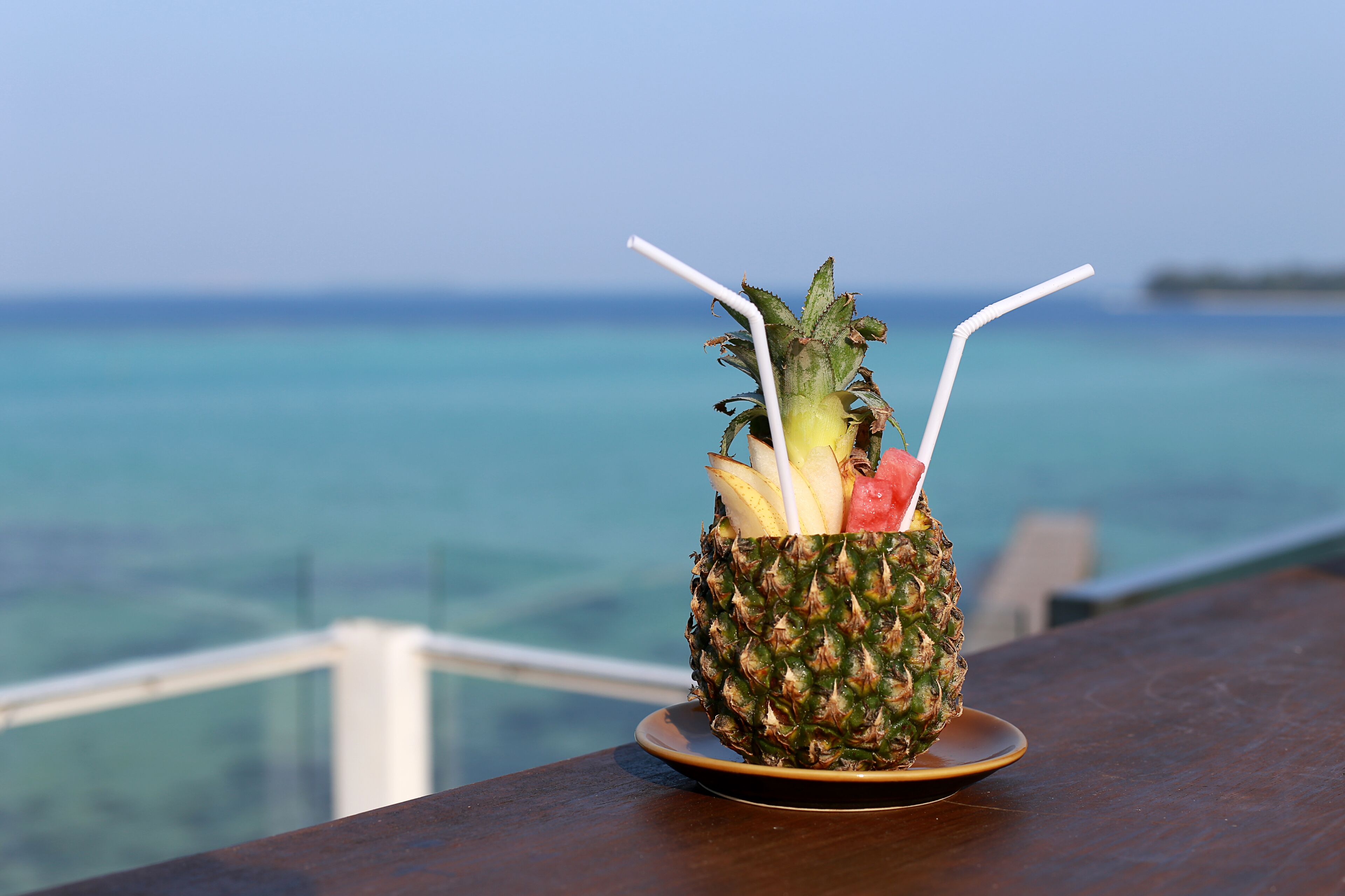 Un ananas avec une paille et une tranche de pastèque sur une assiette, le tout présenté sur fond de vue sur l’océan.