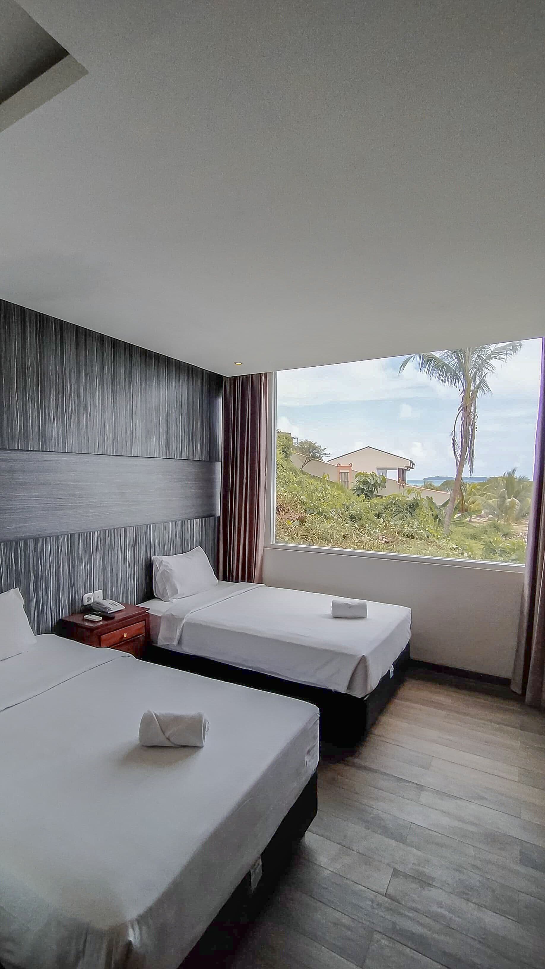 Habitación de hotel con dos camas, cabecero de madera, ventana con vistas a zonas verdes y edificios, y suelo de madera.