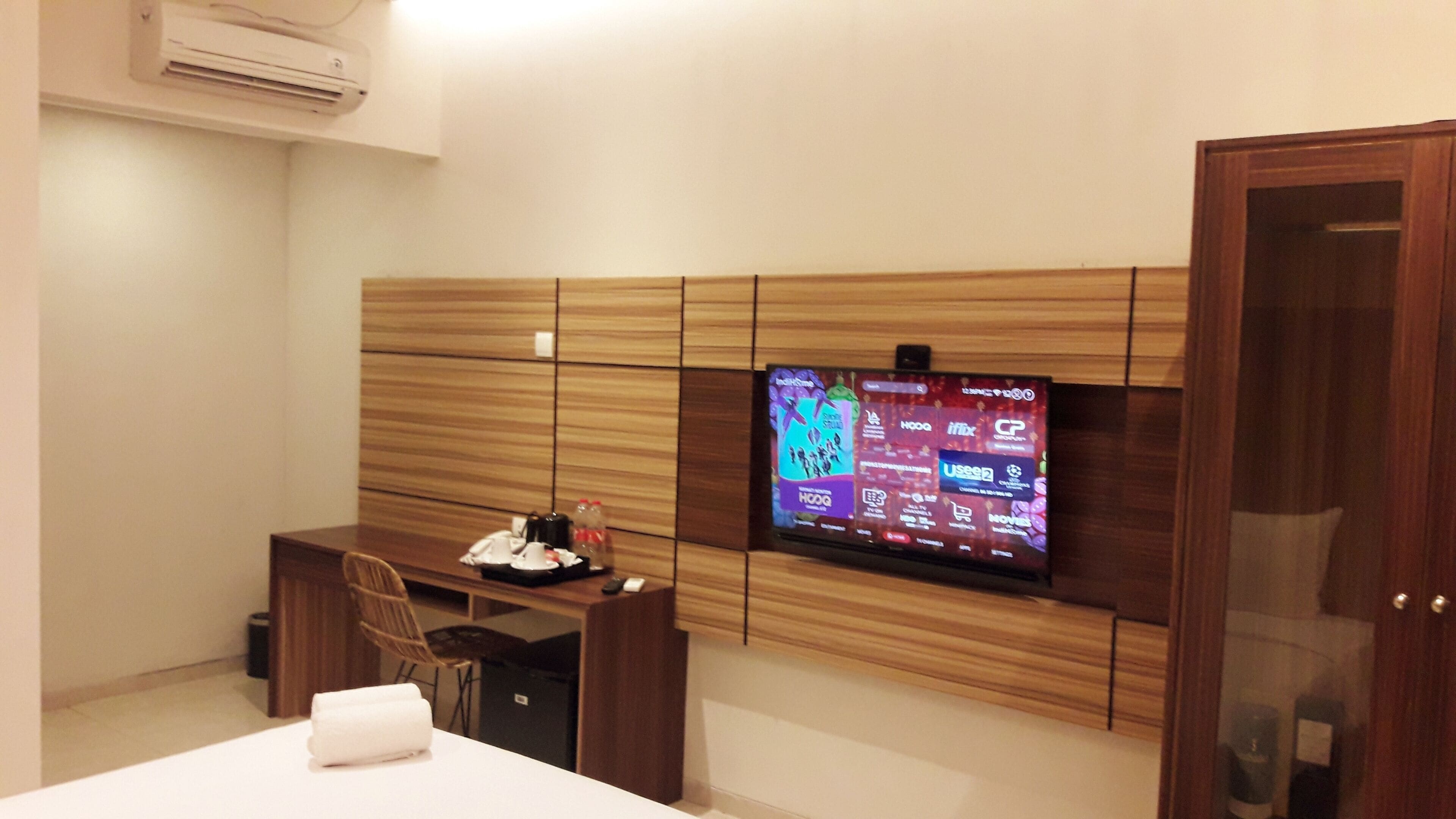 A hotel room with a wooden headboard, a flat-screen TV mounted on the wall, a desk with a chair, and a bed with a white towel.