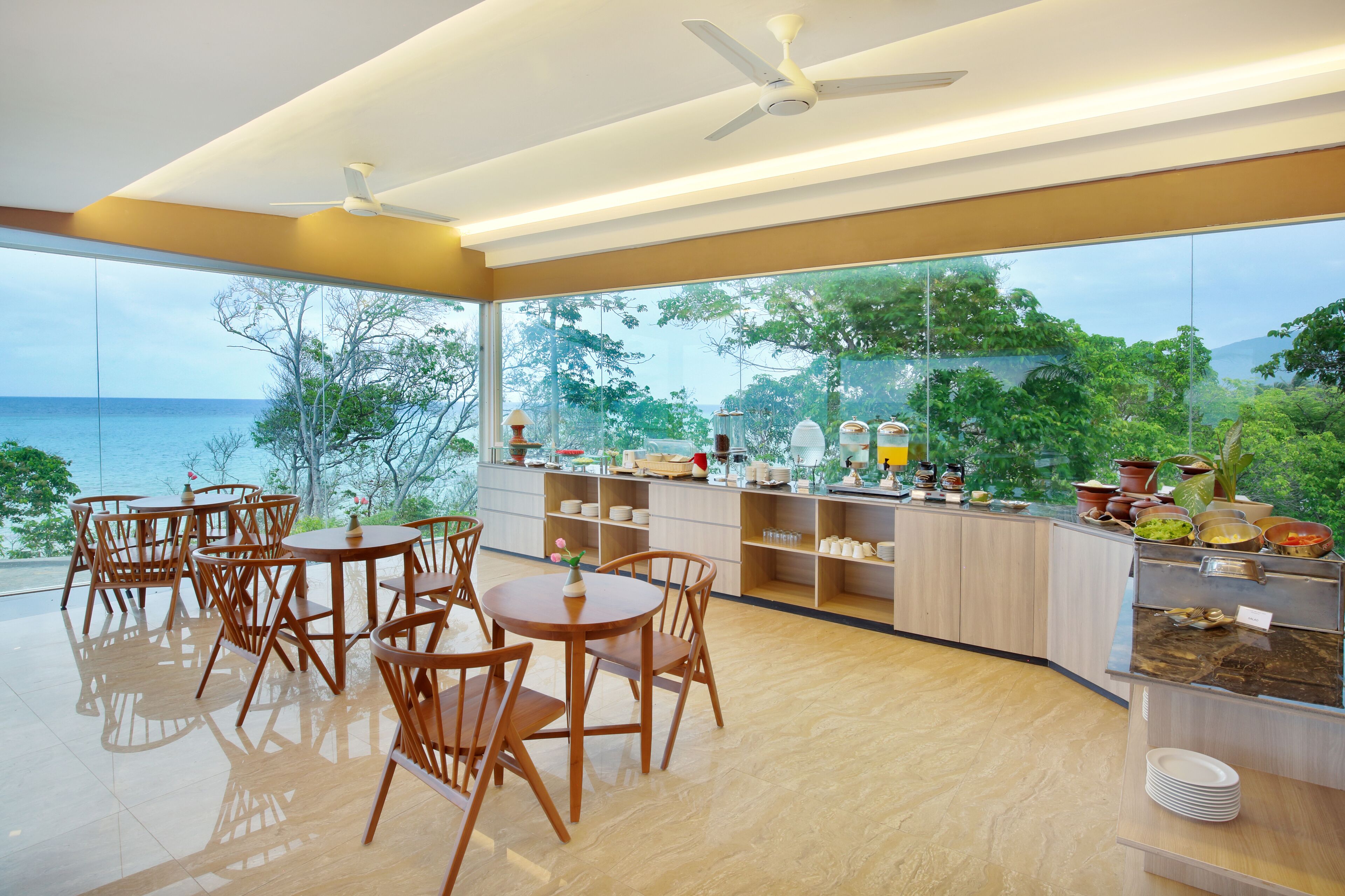 A modern dining area with wooden tables and chairs, a buffet counter with various food items, and a large window offering a view of the sea and greenery.