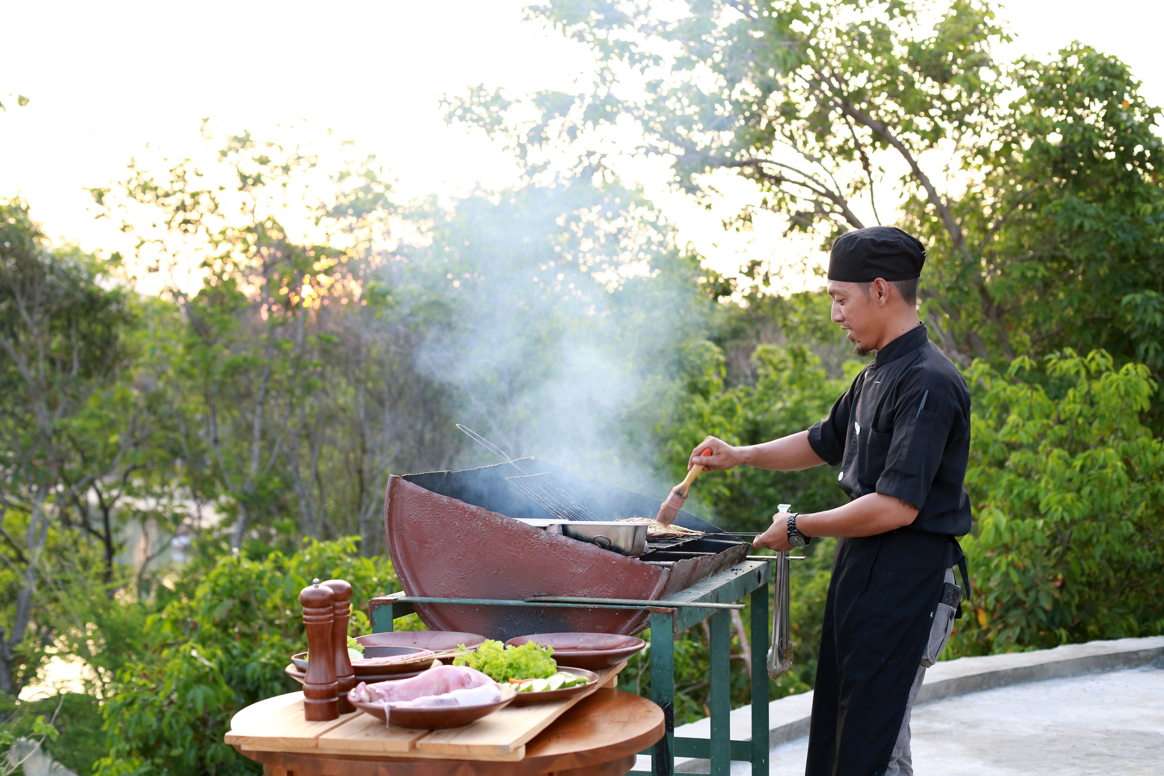 A person grilling outdoors with a barbecue grill and various food items on a table.