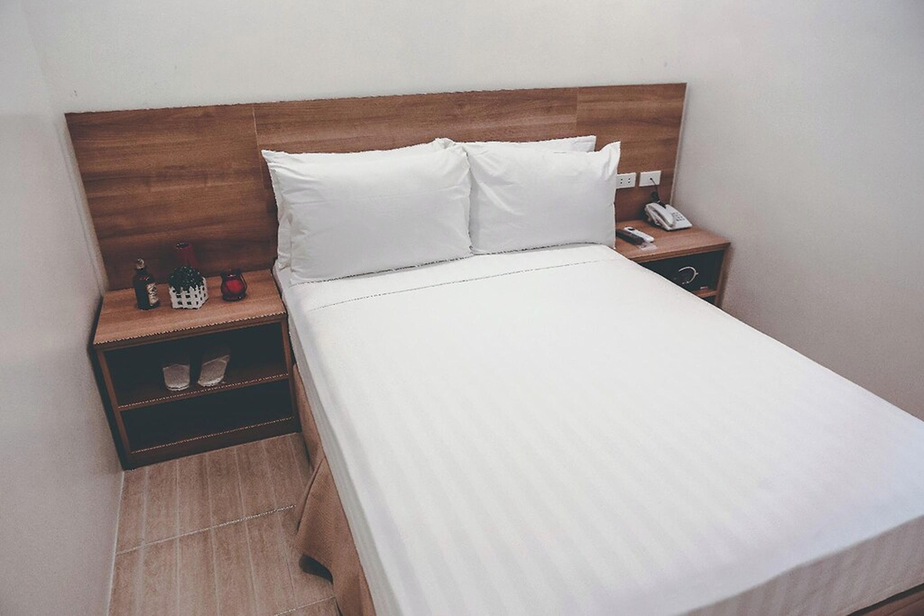 A neatly made bed with white linens and two pillows, a wooden headboard, a bedside table with a phone and a small bottle, and a nightstand with a drawer.