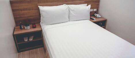 A neatly made bed with white linens and two pillows, a wooden headboard, a bedside table with a phone and a small bottle, and a nightstand with a drawer.