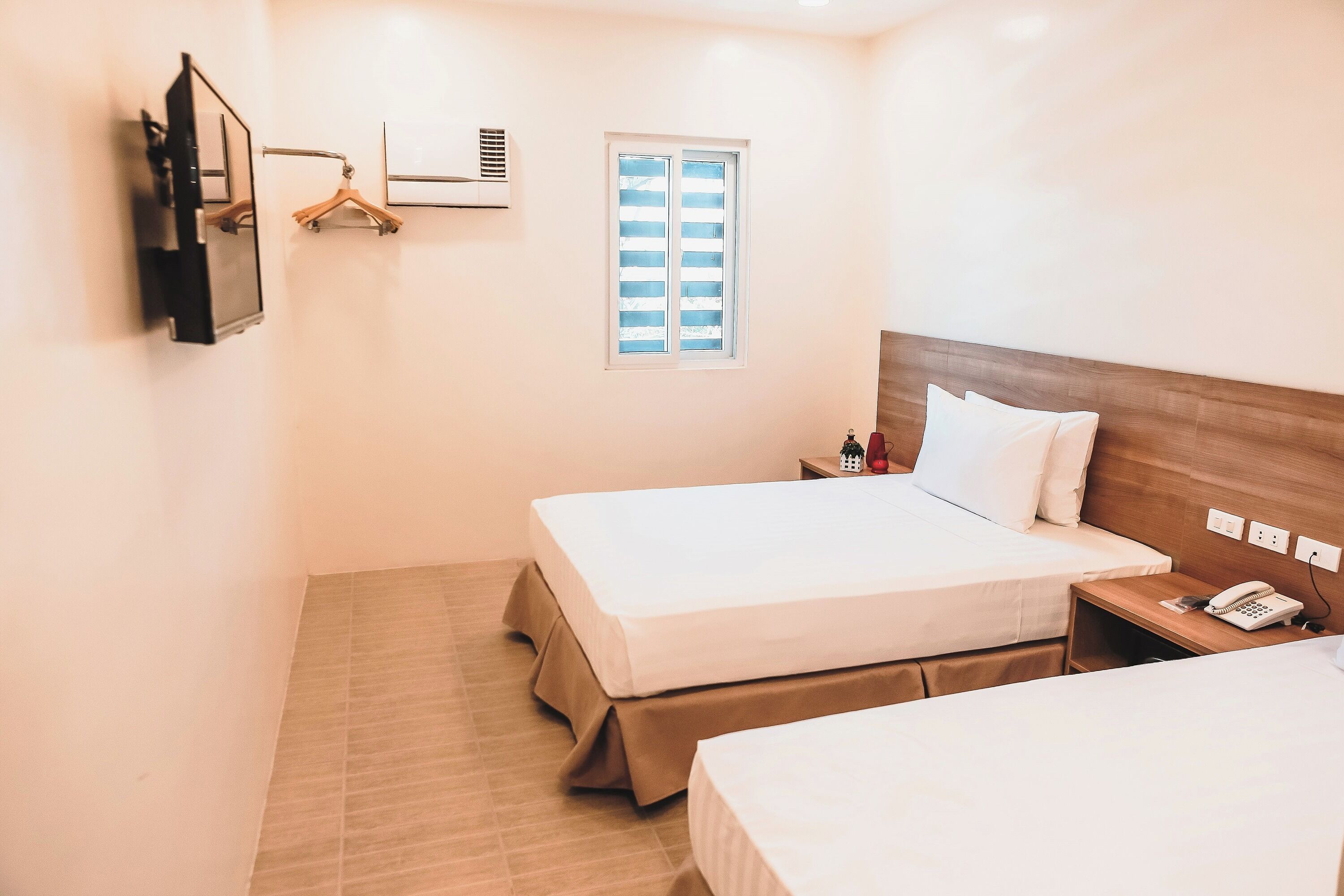 Habitación de hotel con dos camas, cabecera de madera, mesita de noche con teléfono, ventana pequeña y televisor montado en la pared.