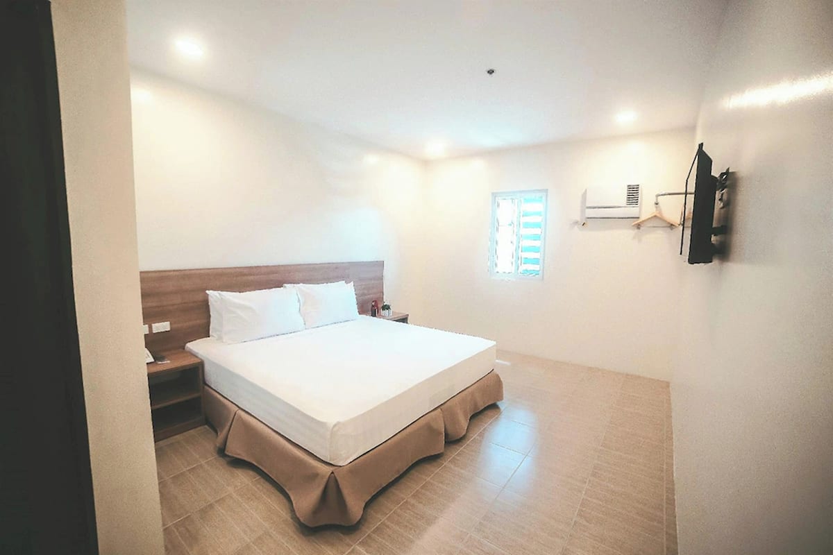 A hotel room with a large bed, a nightstand, a wall-mounted TV, and a window with blinds.