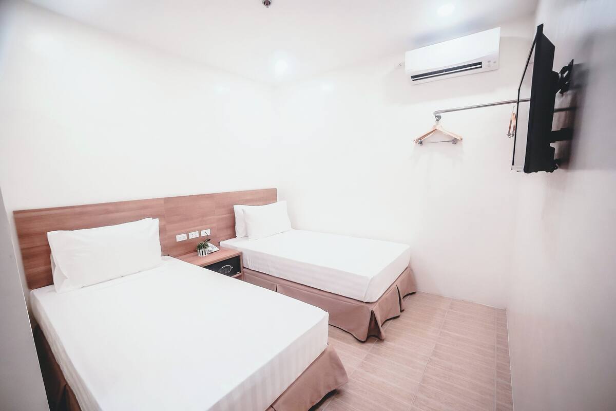 A hotel room with two beds, a wooden headboard, a bedside table, a hanging coat rack, and an air conditioning unit.