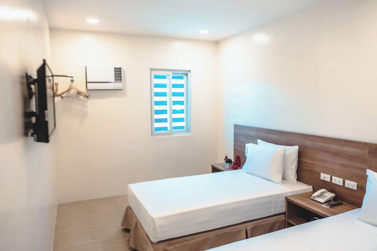 A hotel room with two beds, a wooden headboard, a bedside table with a phone, and a window with blinds.