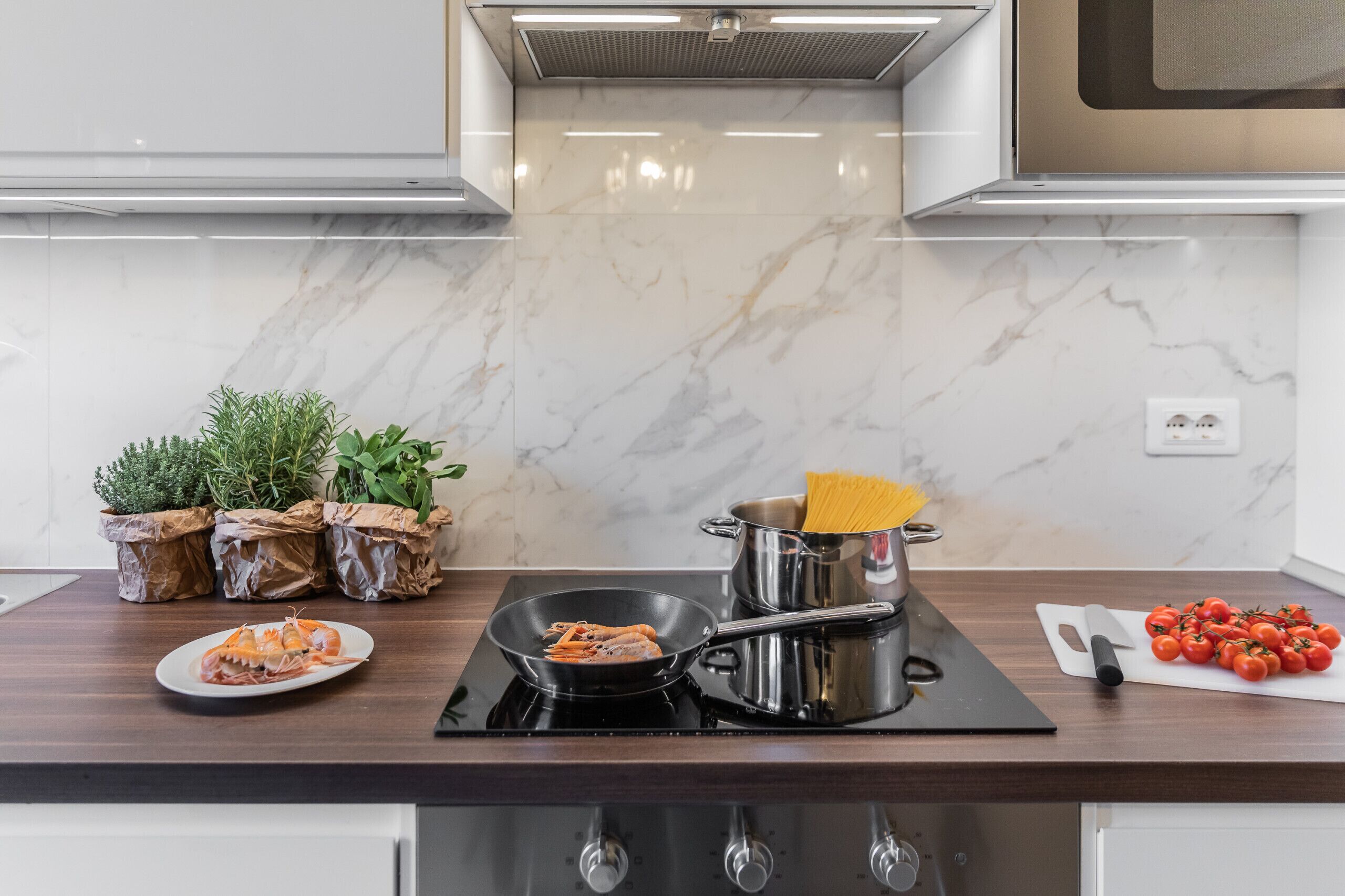 Una cocina con salpicadero de mármol, estufa, ollas, tabla de cortar con tomates y plantas en macetas.