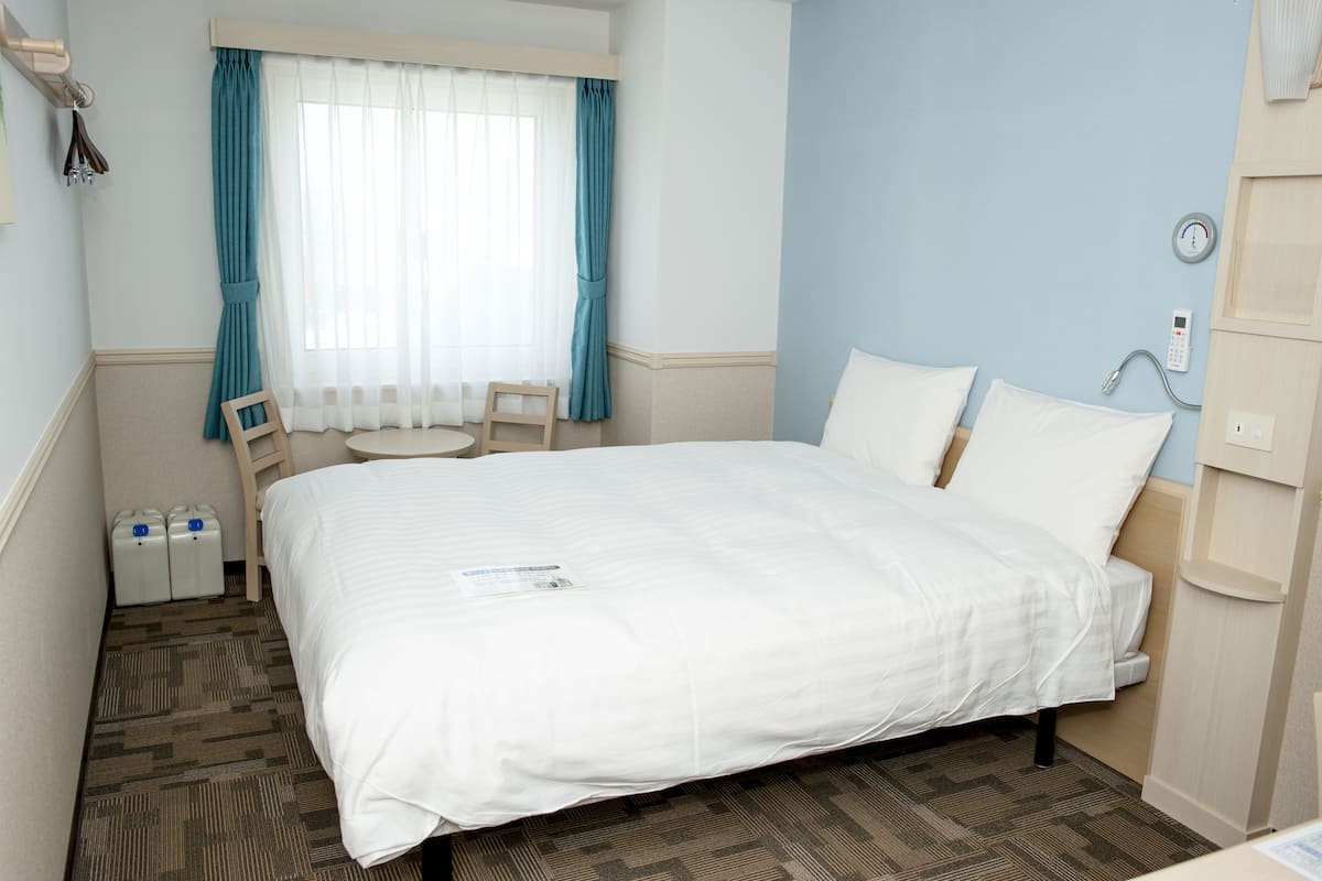 Une chambre d’hôtel comprenant un lit, une chaise, une fenêtre avec des rideaux, une table de chevet et un thermostat.