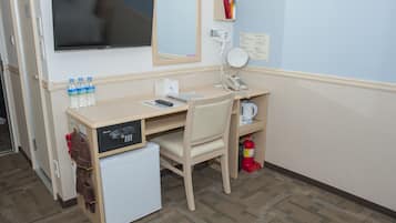 A hotel room with a desk, a television, a mirror, a mini-fridge, a coffee maker, and a fire extinguisher.