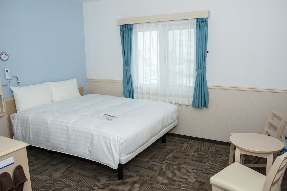 Une chambre d’hôtel avec un lit, une chaise, une table, une horloge et une fenêtre avec des rideaux.