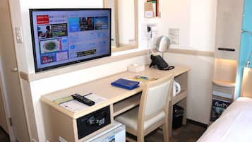 A hotel room with a desk, a television displaying a booking interface, a telephone, and a small trash bin.