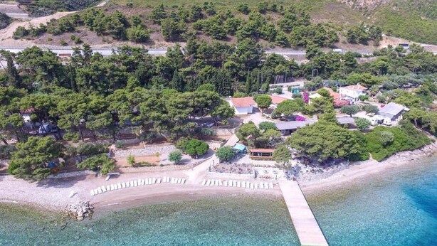 An aerial view of a coastal area with a pier extending into clear blue waters. There are several buildings with visible roofs and structures, surrounded by dense greenery. - Tsolis Village (Aigialeia)