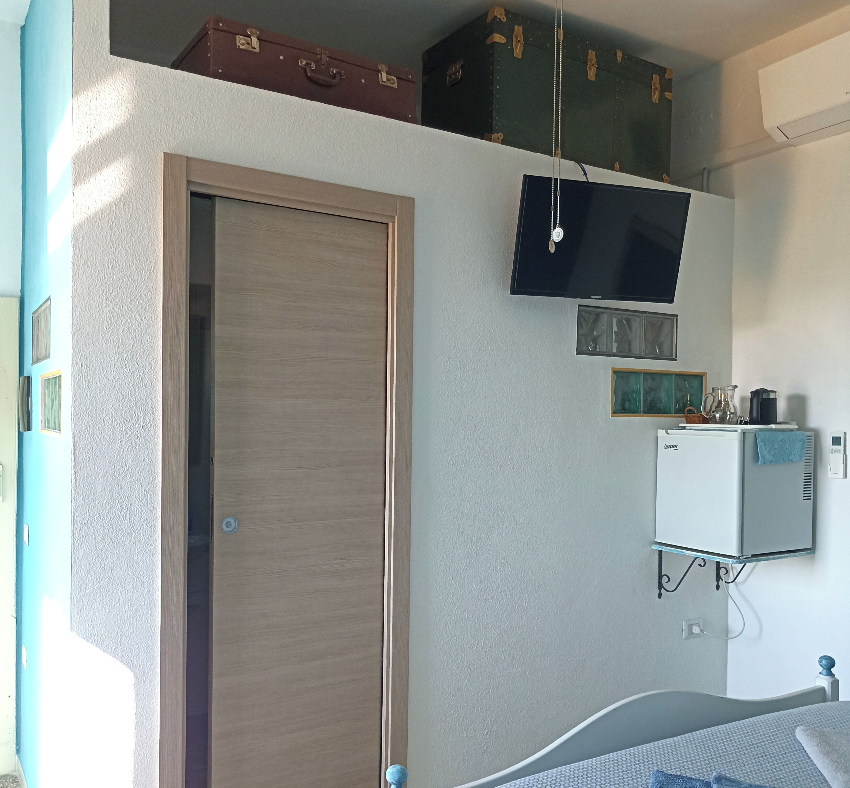 Quarto com porta de madeira, TV de tela plana na parede, pequena geladeira e cama com colcha estampada.