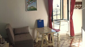 A room with a sofa, a table, a chair, a window with curtains, a wall-mounted picture, and a ceiling light. - B&B Eleonora D'Arborea (Posada)