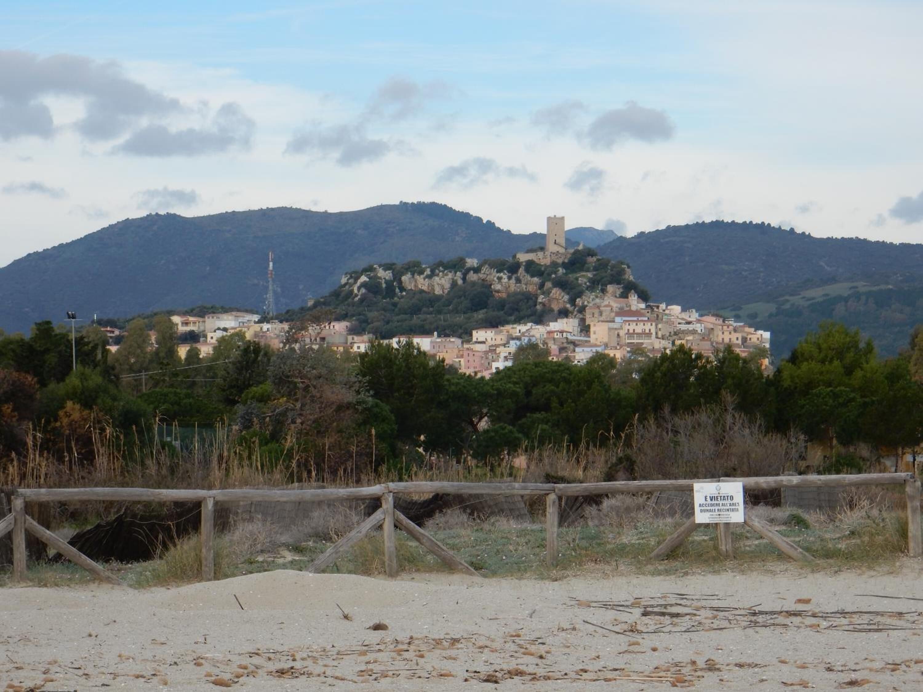 A coastal town with a historic tower on a hill, surrounded by mountains and a sandy foreground.