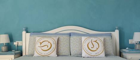 A bedroom with a teal wall, a white bed frame, and two decorative pillows with a circular design. There are two bedside tables with lamps and folded towels on the bed.
