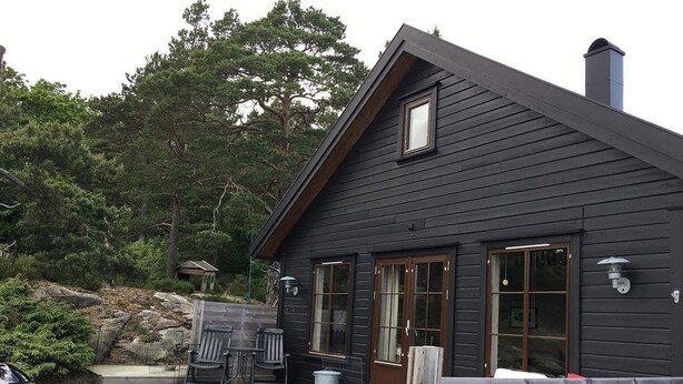 A dark wooden cabin with a sloped roof, large windows, and a deck with outdoor furniture. - Solferie Holiday Cottage Langenes (Kristiansand)