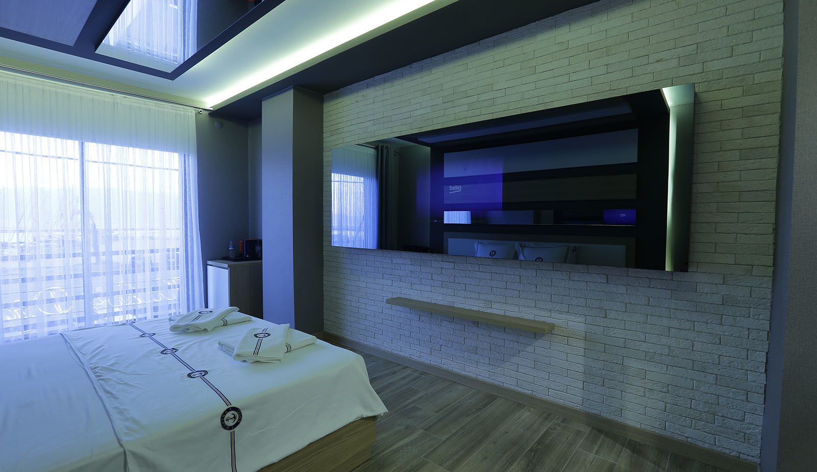 A hotel room with a bed, a TV mounted on a brick wall, and a window with sheer curtains.