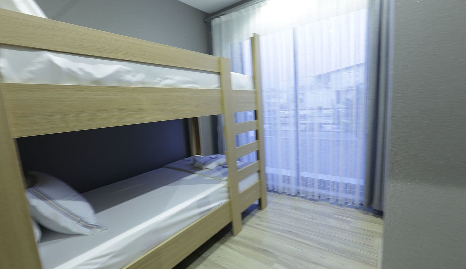 Ein Stockbett mit Matratze und Kissen, eine Holzleiter und ein Fenster mit durchsichtigen Vorhängen.