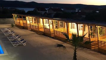 A row of wooden cabins with windows, a pool area with lounge chairs, and a view of hills in the background.