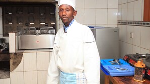 Un chef, vêtu d’un uniforme blanc et d’un tablier à carreaux, se tient dans une cuisine aux murs carrelés et aux appareils en acier inoxydable.