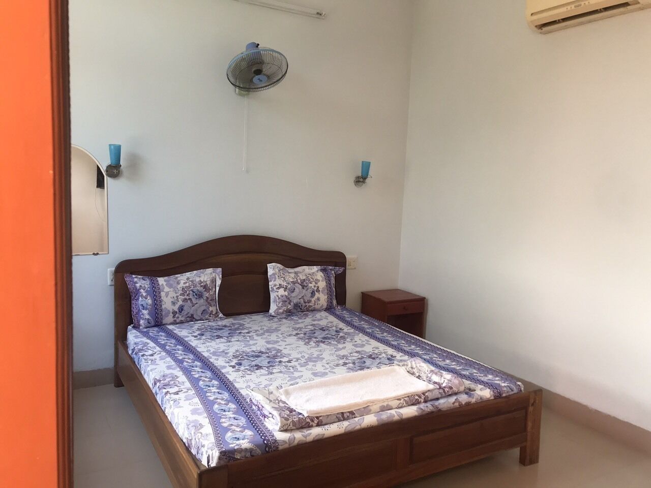 A bedroom with a wooden bed, a bedside table, a wall-mounted fan, and a small nightstand.
