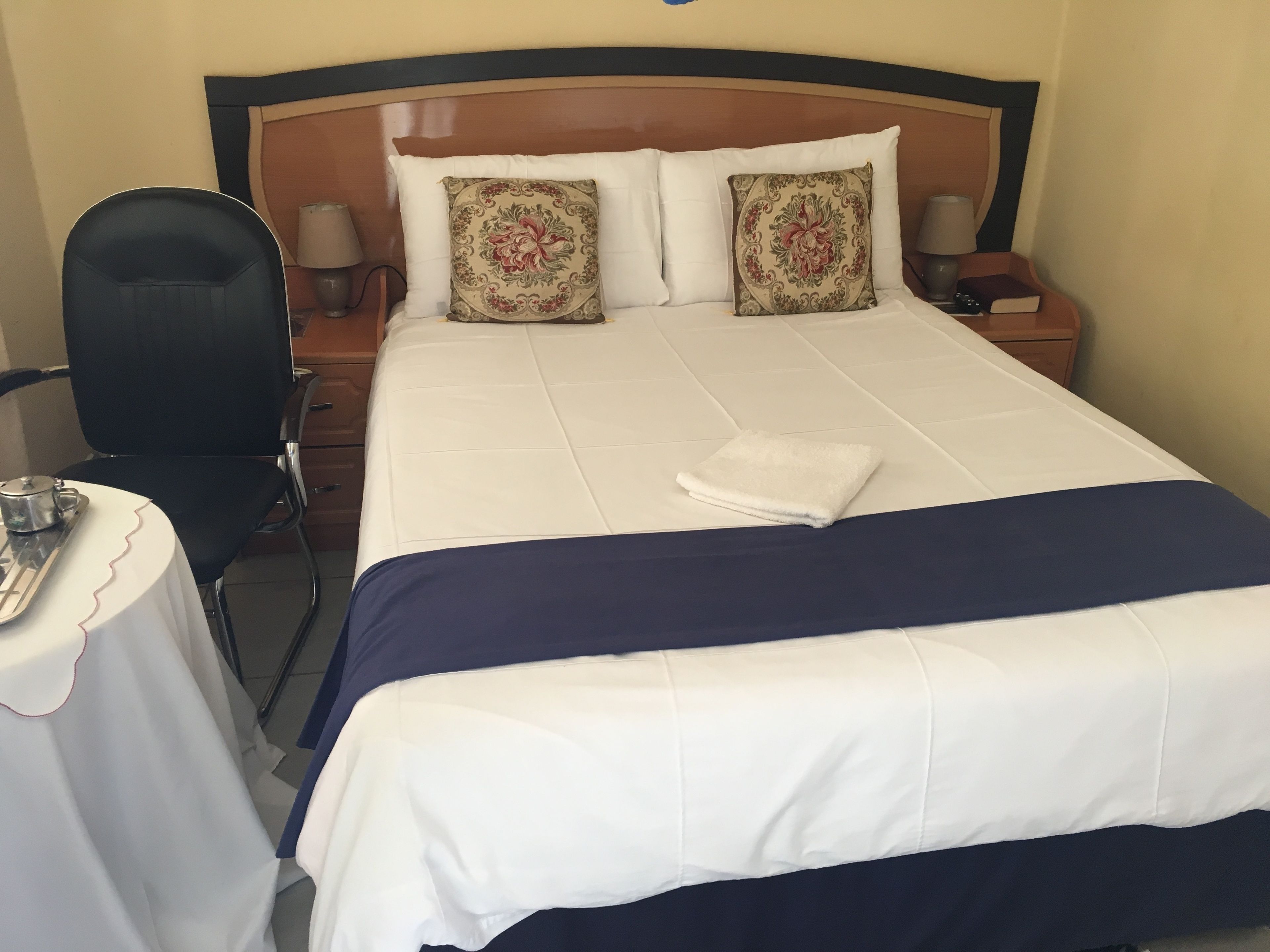 A neatly made bed with a white and navy blue bedspread, two decorative pillows, a folded white towel, a bedside table with a lamp, and a chair.