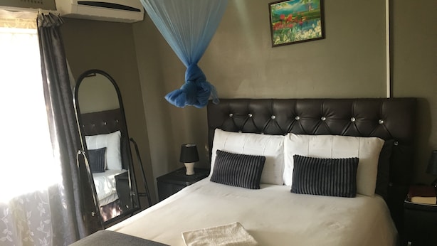 A hotel room with a large bed, a nightstand, a lamp, a mirror, and a window with curtains. - Kutse Guesthouse (Letlhakeng)