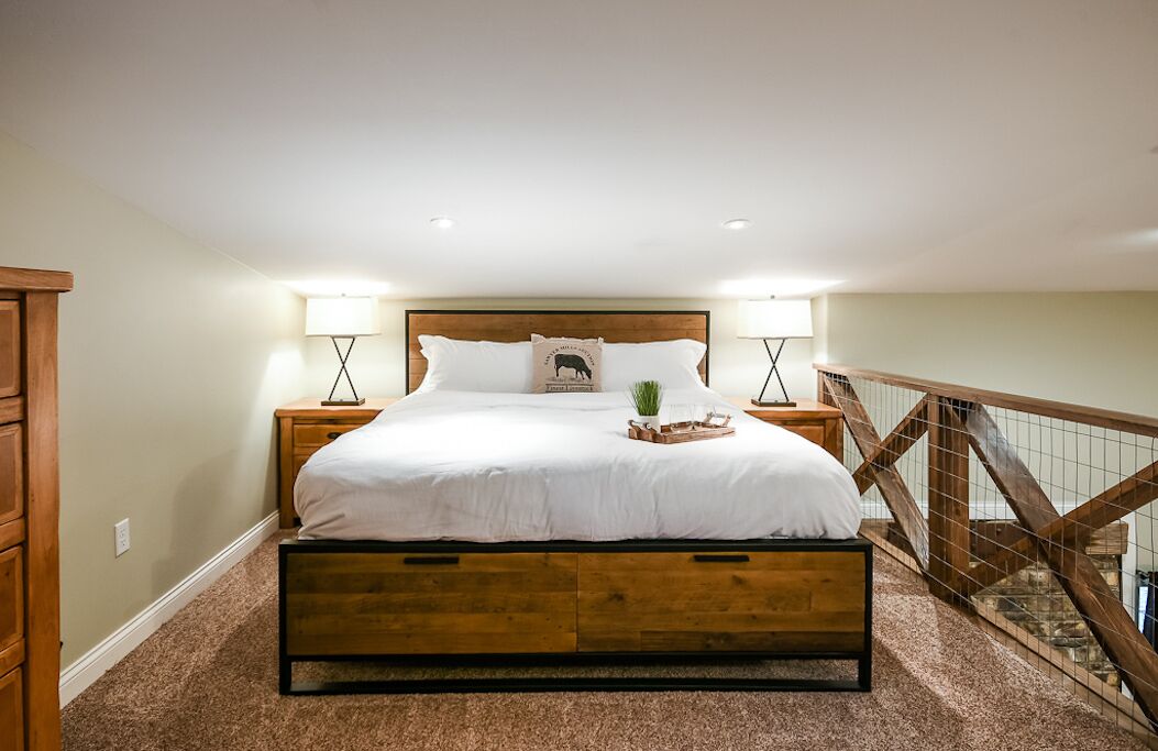 Een slaapkamer met een houten bed, twee nachtkastjes, een bureautje, een lamp, een decoratief kussen en een kleine plant.