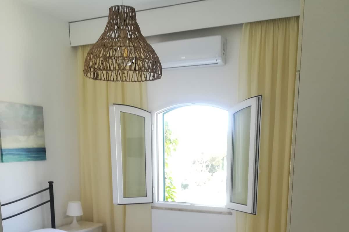A bedroom with a bed, a bedside table, a lamp, a window with curtains, and a hanging light fixture.