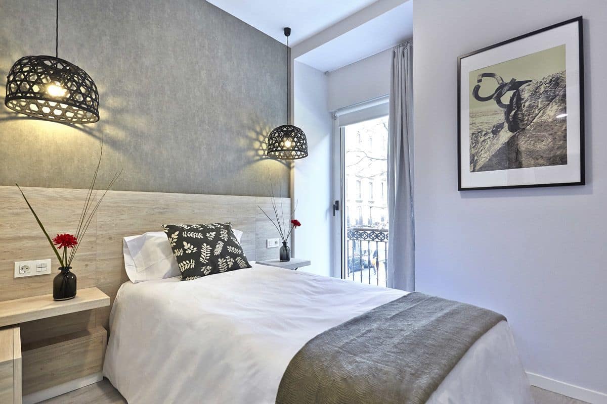 A modern bedroom with a large bed, two pendant lights, a framed artwork, and a view of the outdoors.