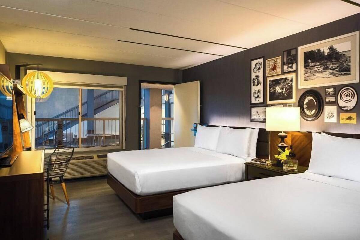 A hotel room with two beds, a desk, a chair, a lamp, and a wall with framed pictures.