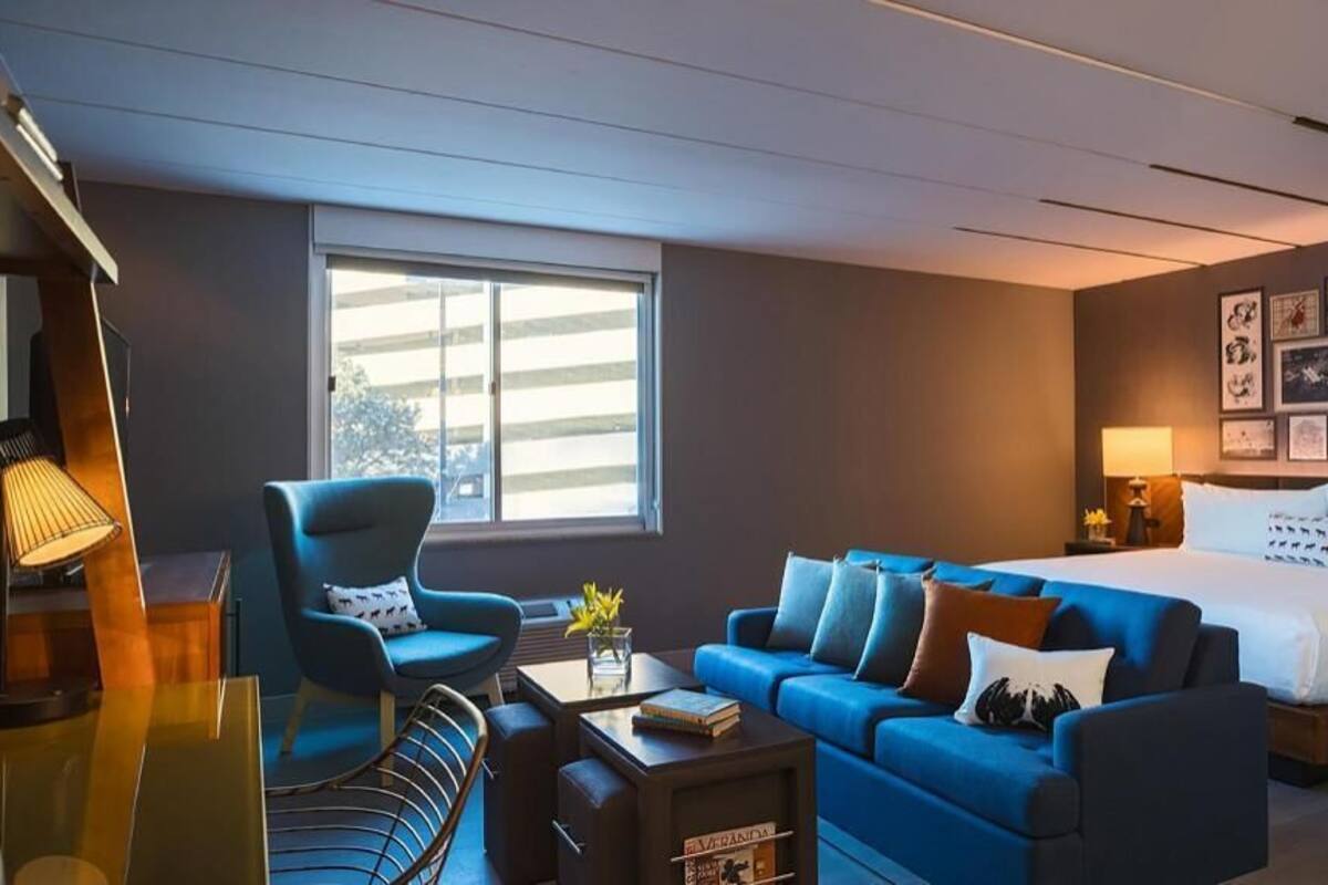 A hotel room with a bed, a blue sofa, a desk with a lamp, and a window.