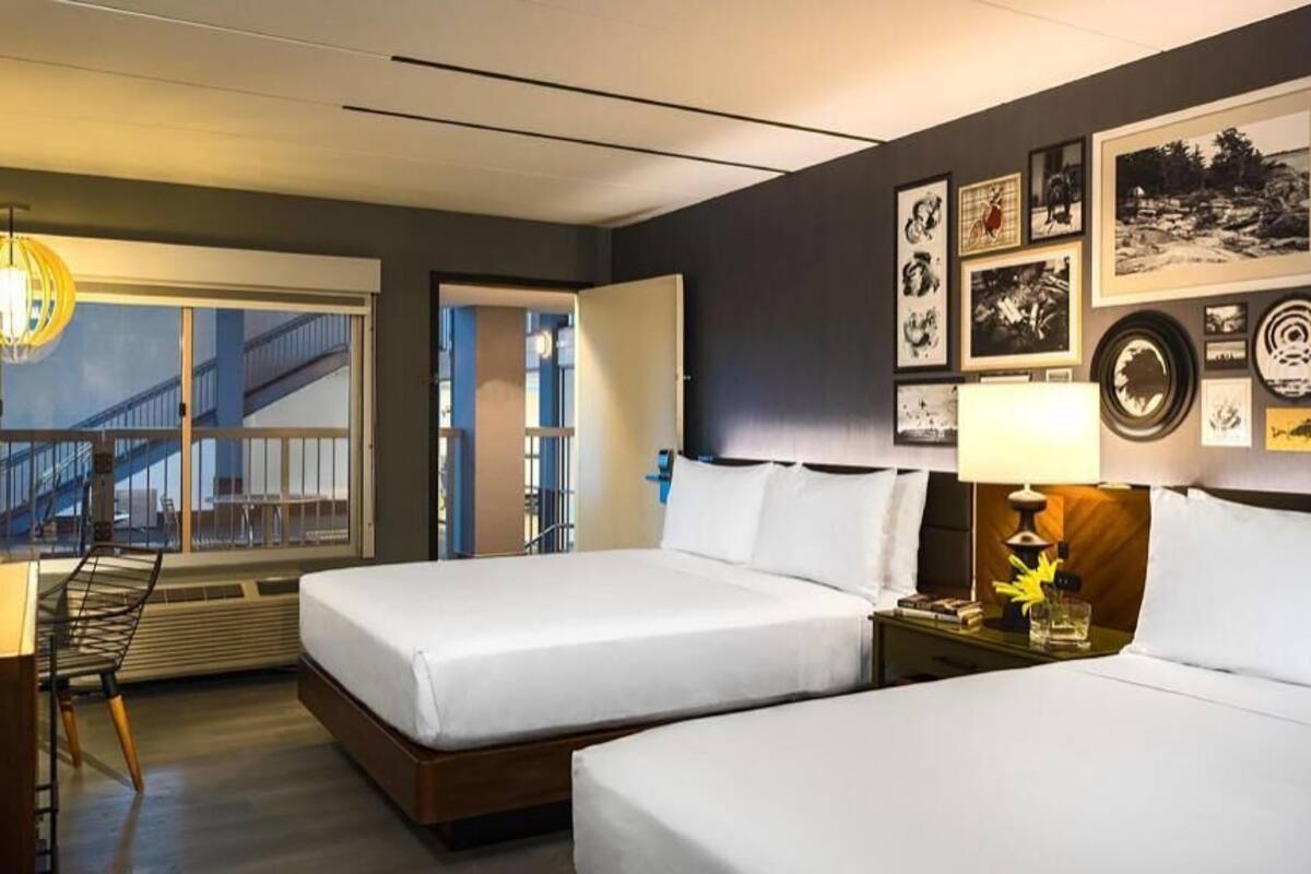 A hotel room with two beds, a desk, a chair, a lamp, and a wall with framed pictures.