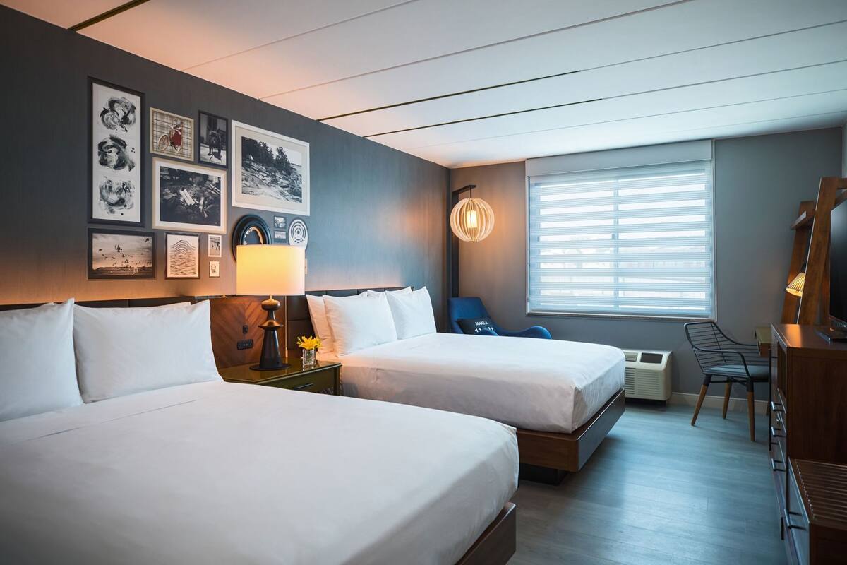 A hotel room with two beds, a desk, a chair, a TV, and artwork on the wall.