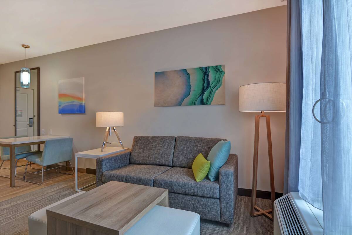 Une chambre d’hôtel moderne avec un canapé gris, une petite table, une lampe et un tableau au mur.