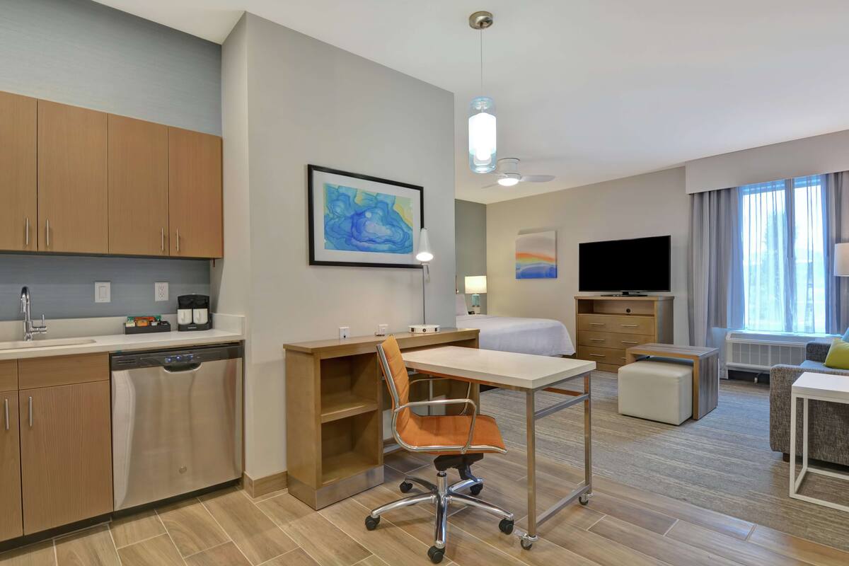 Une chambre d’hôtel moderne dotée d’une kitchenette, d’un coin repas, d’une télévision et d’une grande fenêtre avec des rideaux.