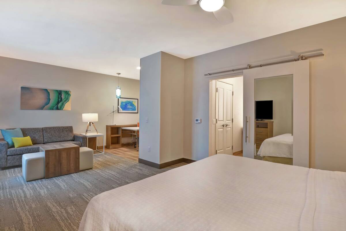 Une chambre d’hôtel avec un grand lit, un canap é, un bureau et une chaise. Une porte donne sur une autre pièce et il y a un téléviseur dans un coin.