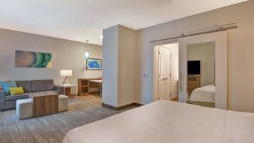 Ein Hotelzimmer mit einem großen Bett, einer Couch, einem Schreibtisch und einem Sessel. Eine Tür führt in einen weiteren Raum, und in der Ecke befindet sich ein Fernseher.