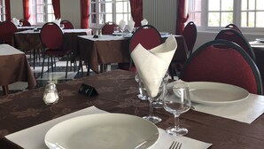 A dining area with tables set for two, featuring white plates, silverware, and clear glassware. Red curtains and windows with blinds are visible. - Hôtel Restaurant La Diligence (Le Brignon)