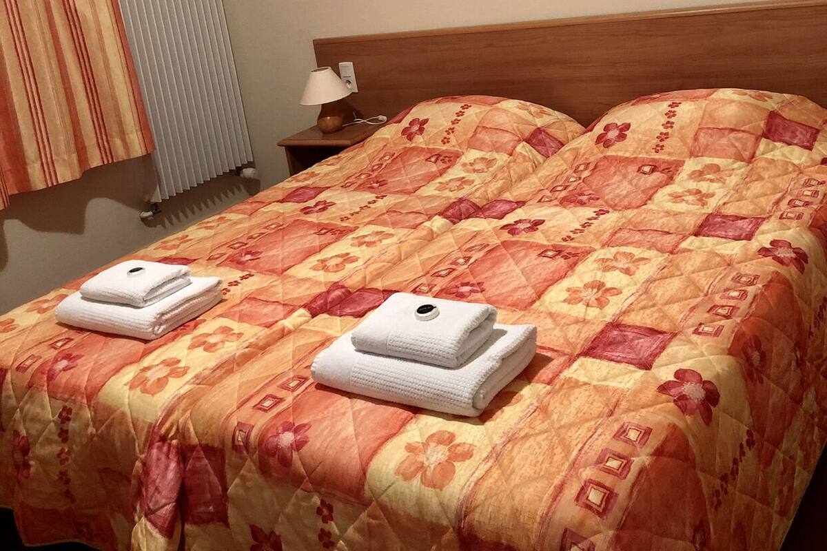 Habitación de hotel con una cama grande, dos almohadas, una mesita de noche con lámpara y una ventana con persianas.