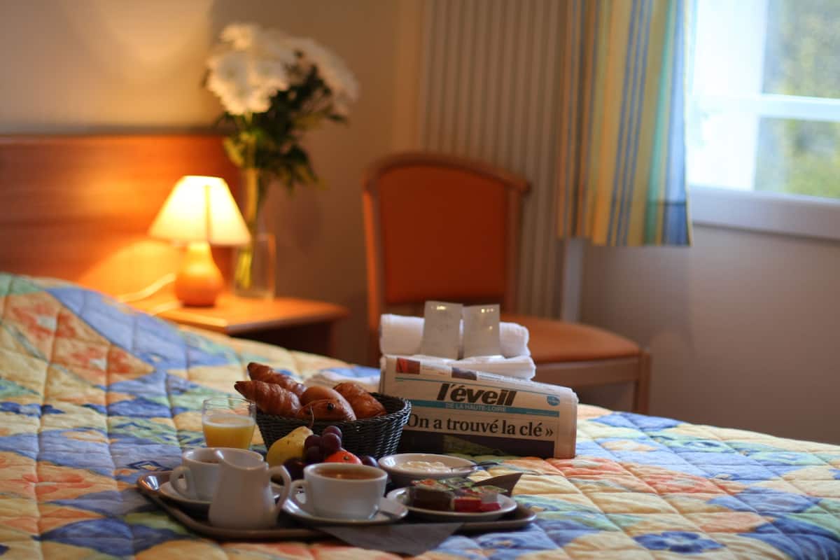 Una bandeja de desayuno con croissants, fruta y bebidas sobre una cama con colcha floral. También se ve una caja de café L'evell y un jarrón con flores.