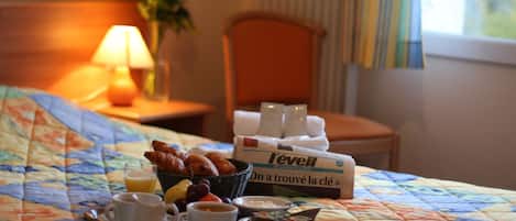 A neatly made breakfast tray with croissants, fruit, and beverages on a bed with a floral quilt. A box of L'evell coffee and a vase of flowers are also visible.