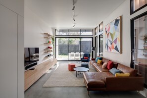 A modern living room with a sofa, coffee table, and TV. There are shelves with decorative items and a large window providing natural light.