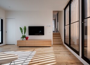 A modern living room with a flat-screen TV mounted on a wall, a wooden entertainment unit, a large window, and a staircase leading to another level.