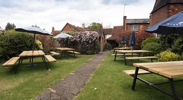 An outdoor patio area with picnic tables and umbrellas, surrounded by brick buildings and flowering bushes.