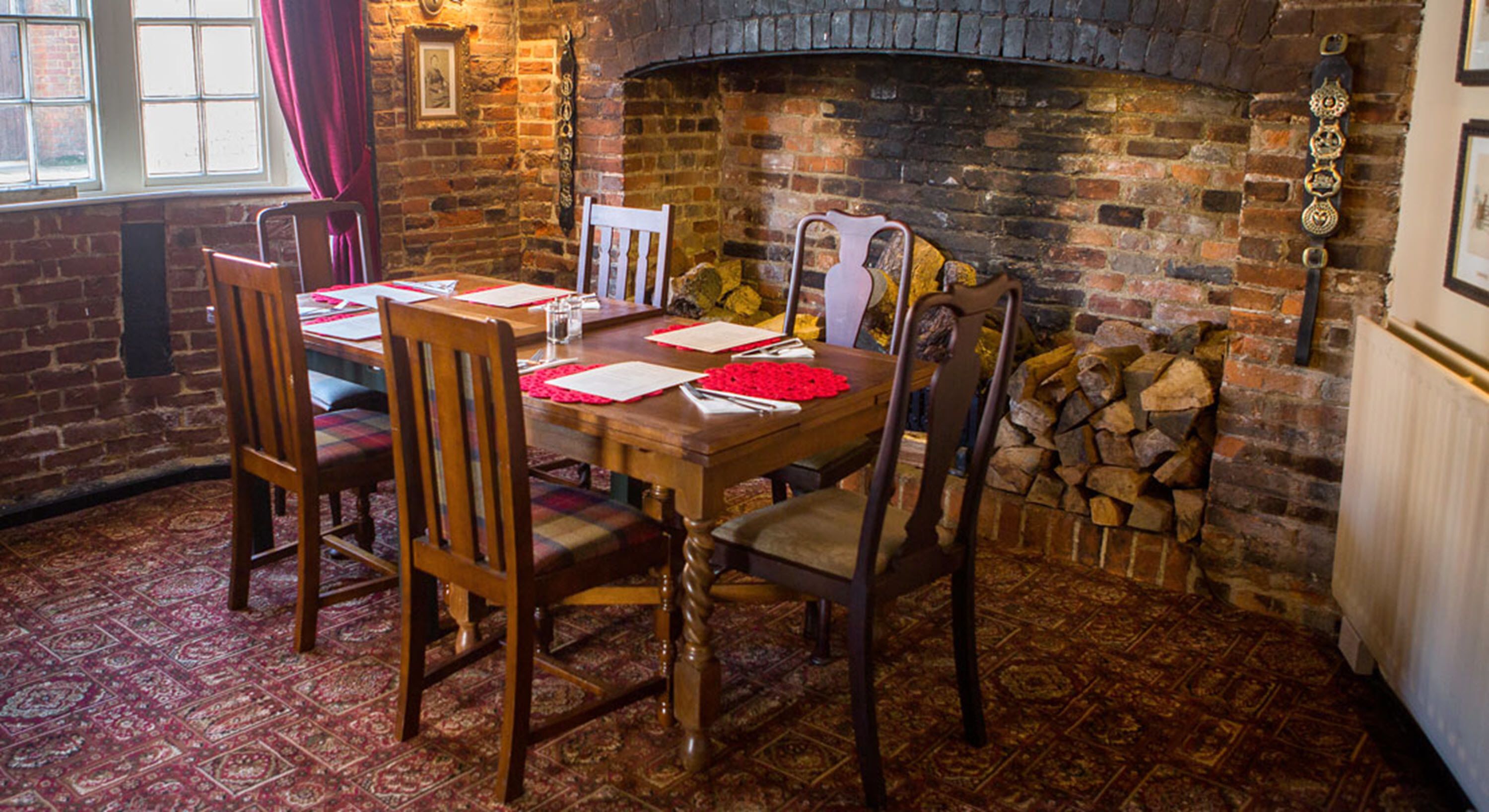 A dining area with a brick fireplace, wooden chairs, a table set with red placemats, and a patterned carpet.