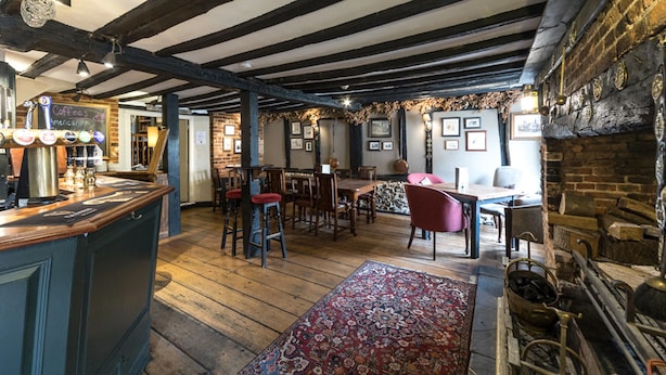 A rustic pub with exposed wooden beams, a bar area with stools, and a traditional fireplace. - The Bull at Wargrave (Reading)